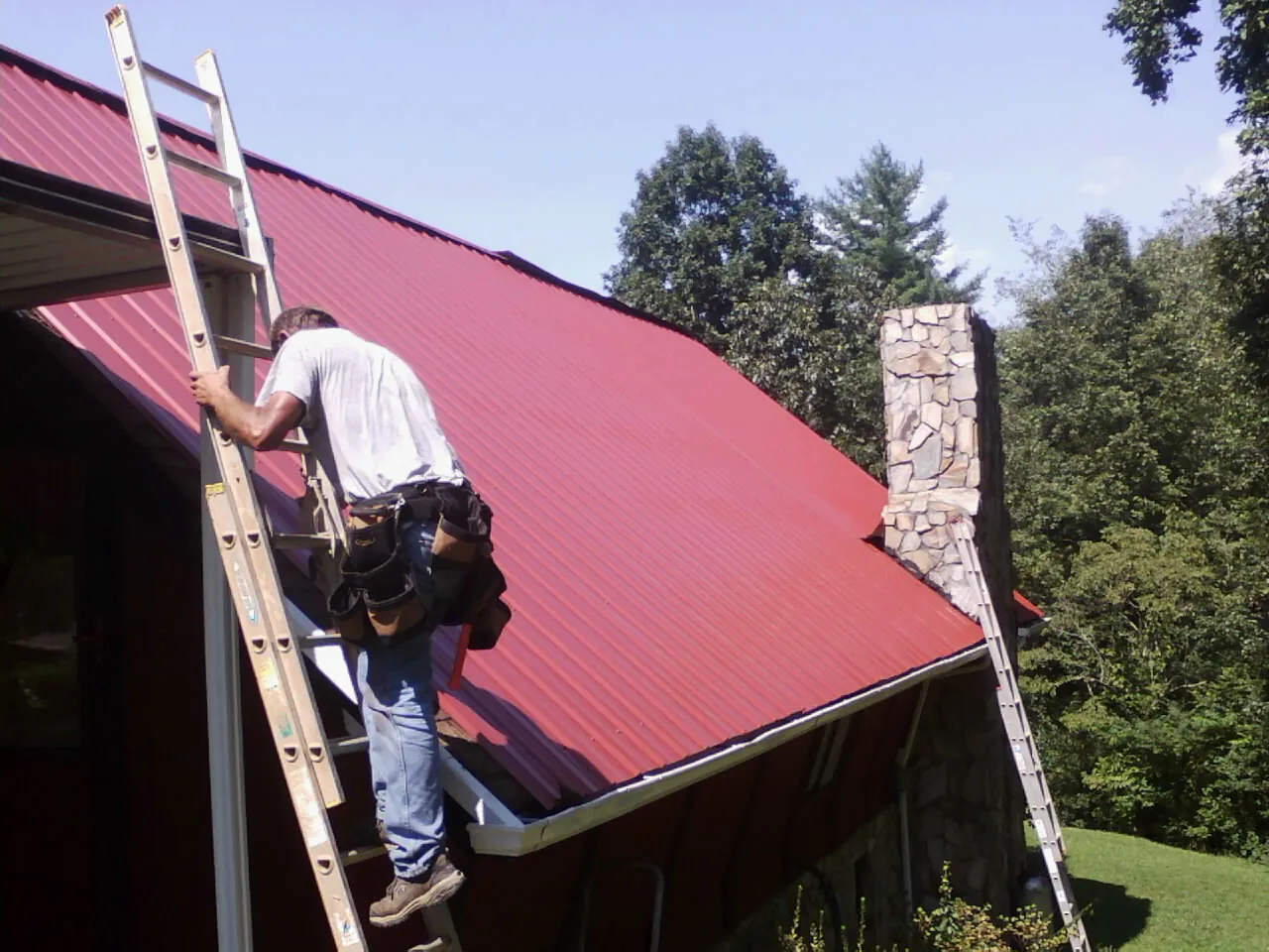 Roofer on a ladder beside a red metal roof and stone chimney — Soffit & Fascia Repair by Roofing Brothers in Upland